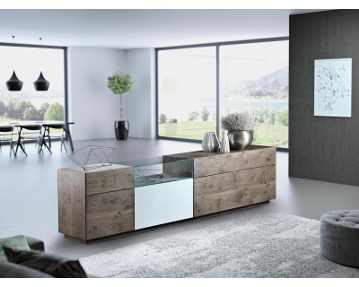 Ein modernes Wohnzimmer mit dem Sideboard „Wohnkombination Sona“, geschmückt mit dekorativen Gegenständen, großen Fenstern und einer malerischen Aussicht auf Berge und einen See.