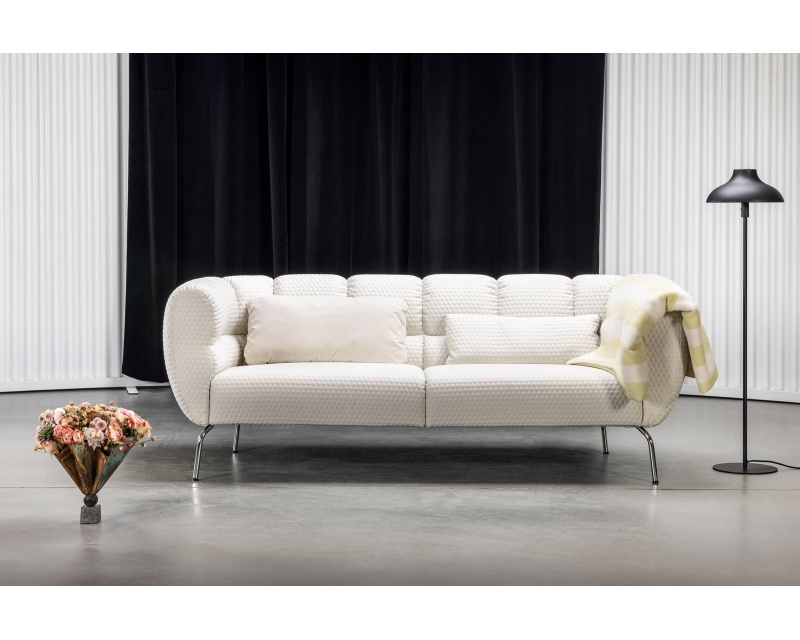 Ein Sofa Magnolia in Weiß mit Kissen und einer Decke steht neben einer schwarzen Stehlampe und einer Vase mit Blumen auf dem Boden.